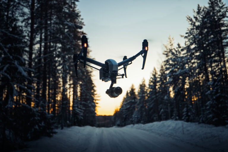 Kamerakopteri leijuu lumisen metsätien yllä auringonlaskussa