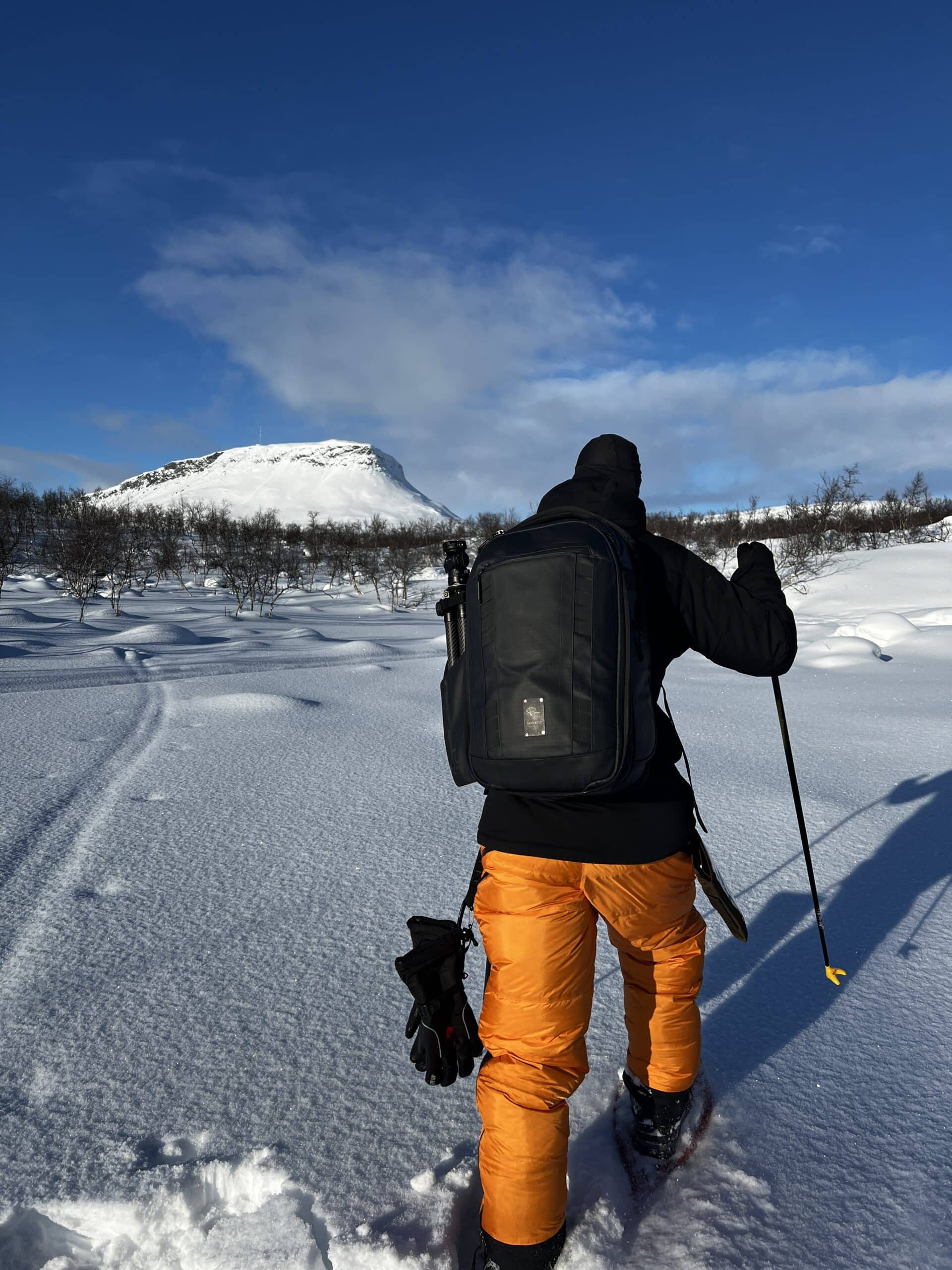 Fraymful crew member on snowshoes crossing a snowfield towards a flat-topped fell, a camera backpack and tripod on the back