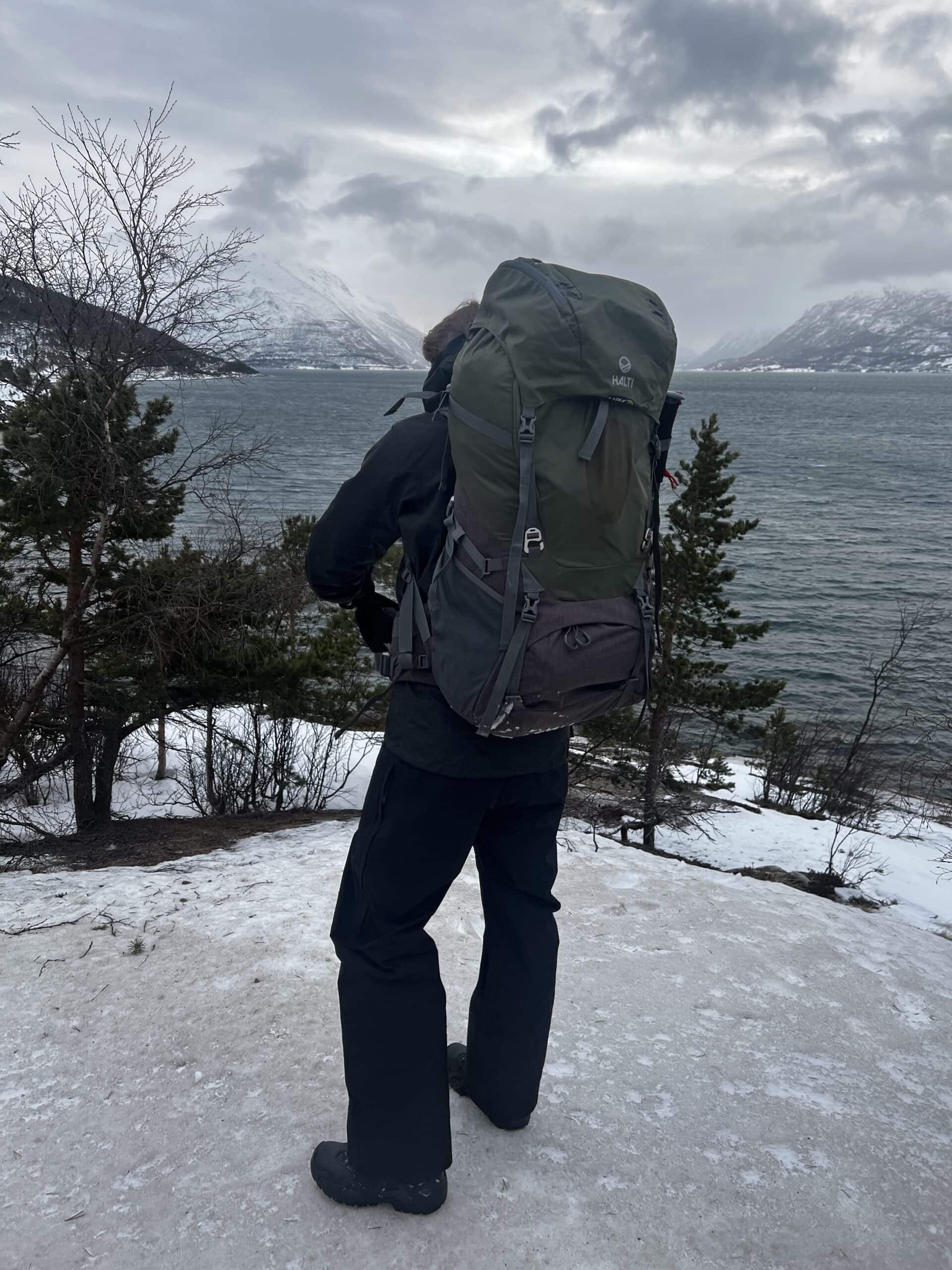 Fraymful crew member with a large expedition rucksack standing on the snow above a fjord, snow-covered mountains across the water