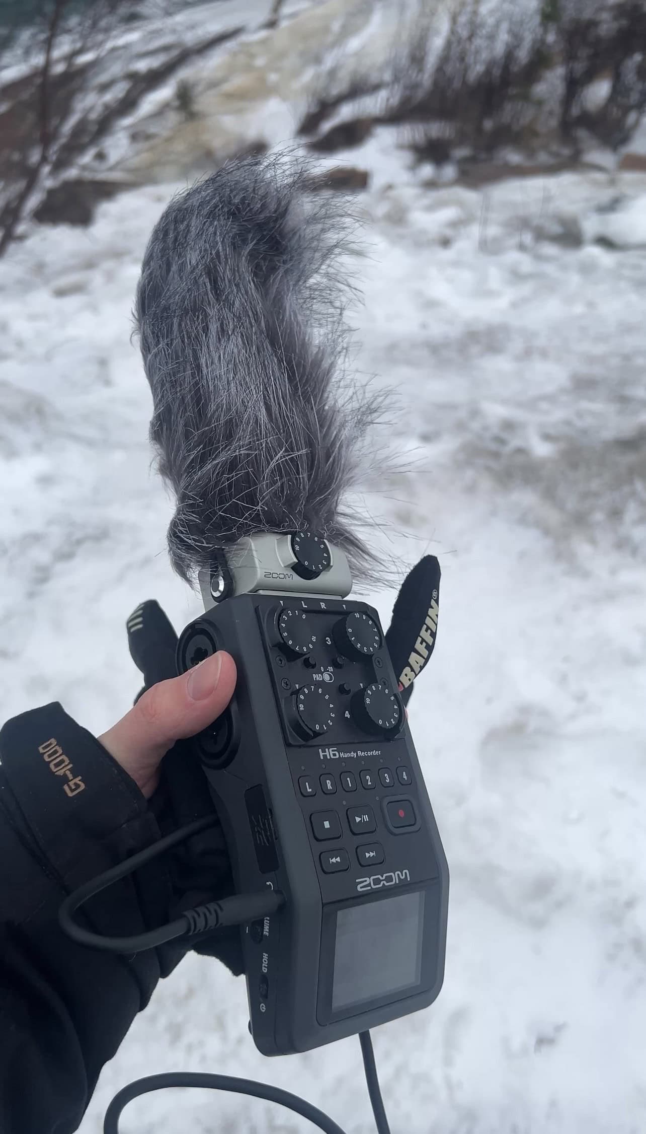Gloved hand holding a Zoom H6 recorder with a furry windshield on the microphone, snowy riverbank behind