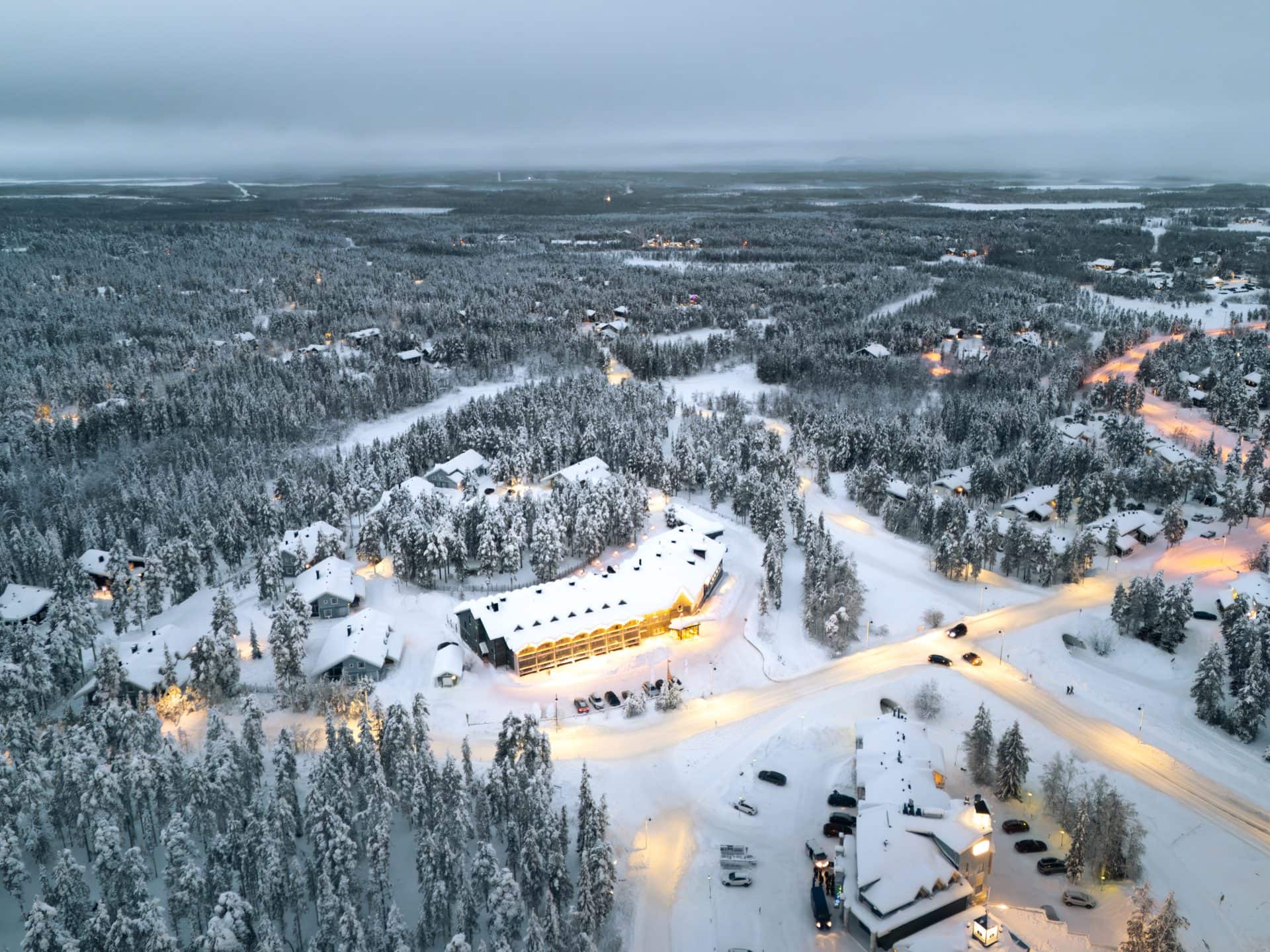Ilmakuva valaistusta Hotel K5 Levistä hämärässä, ympärillä lumisia mökkejä ja mäntyjä Levin kylässä
