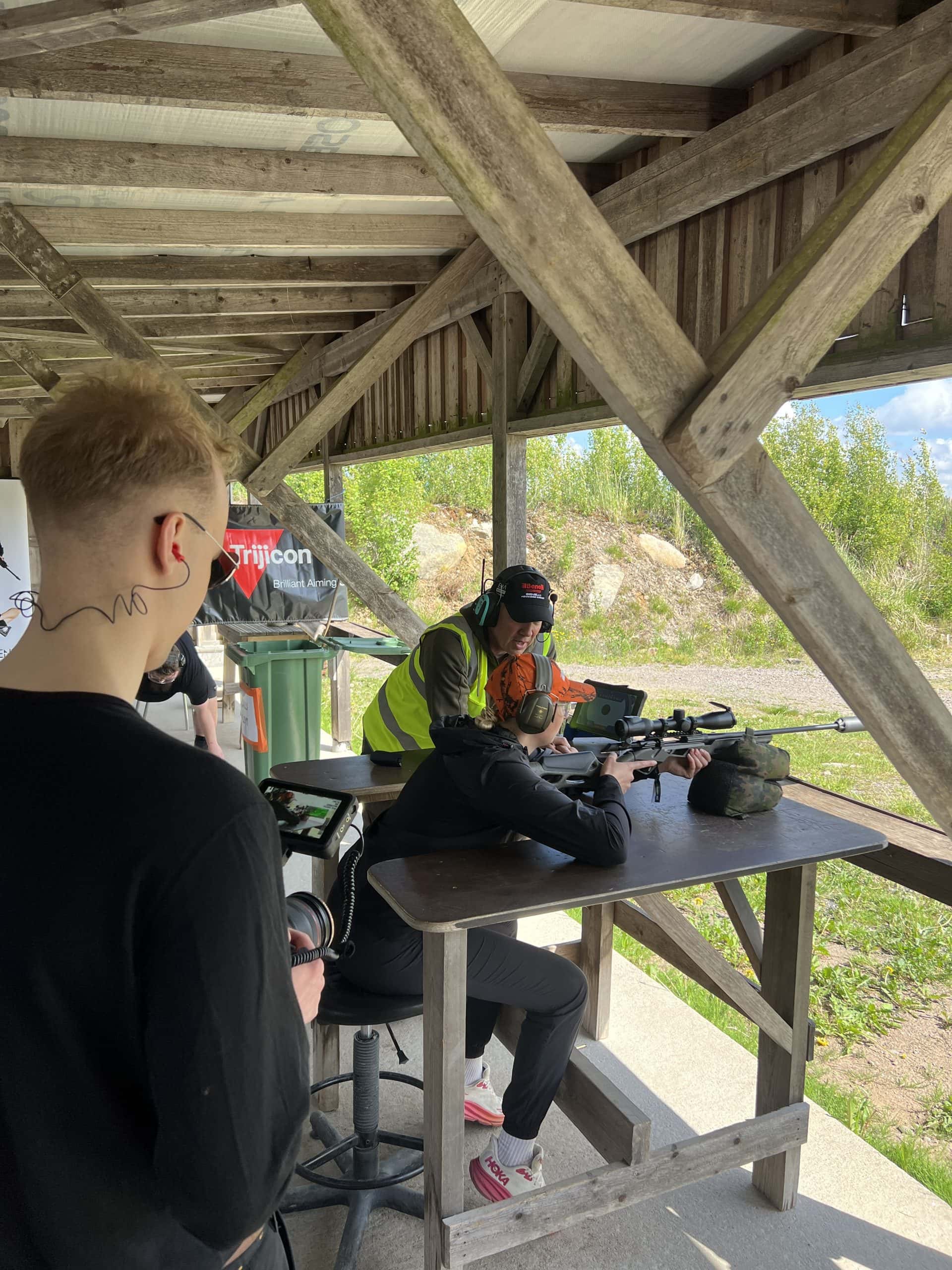 Fraymfulin kuvaaja kuvaa näyttelijää, joka ampuu Benelli Lupo Alpha -kiväärillä tuen päältä kivääripisteellä, ratavalvoja neuvoo takaa.