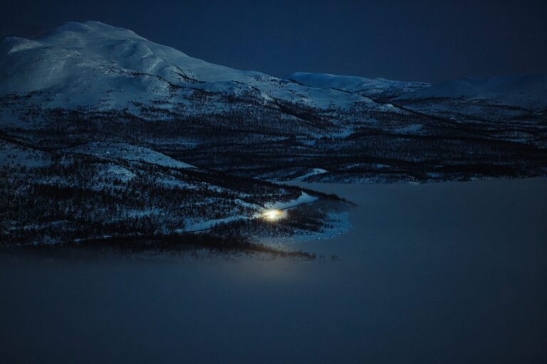 Luminen tunturi jäätyneen järven yllä yöllä Kilpisjärvellä, rannalla hehkuu yksi valo