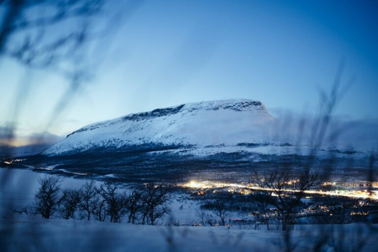 Laakea luminen tunturi Kilpisjärven kylän valojen yllä sinisenä hetkenä, etualalla oksia